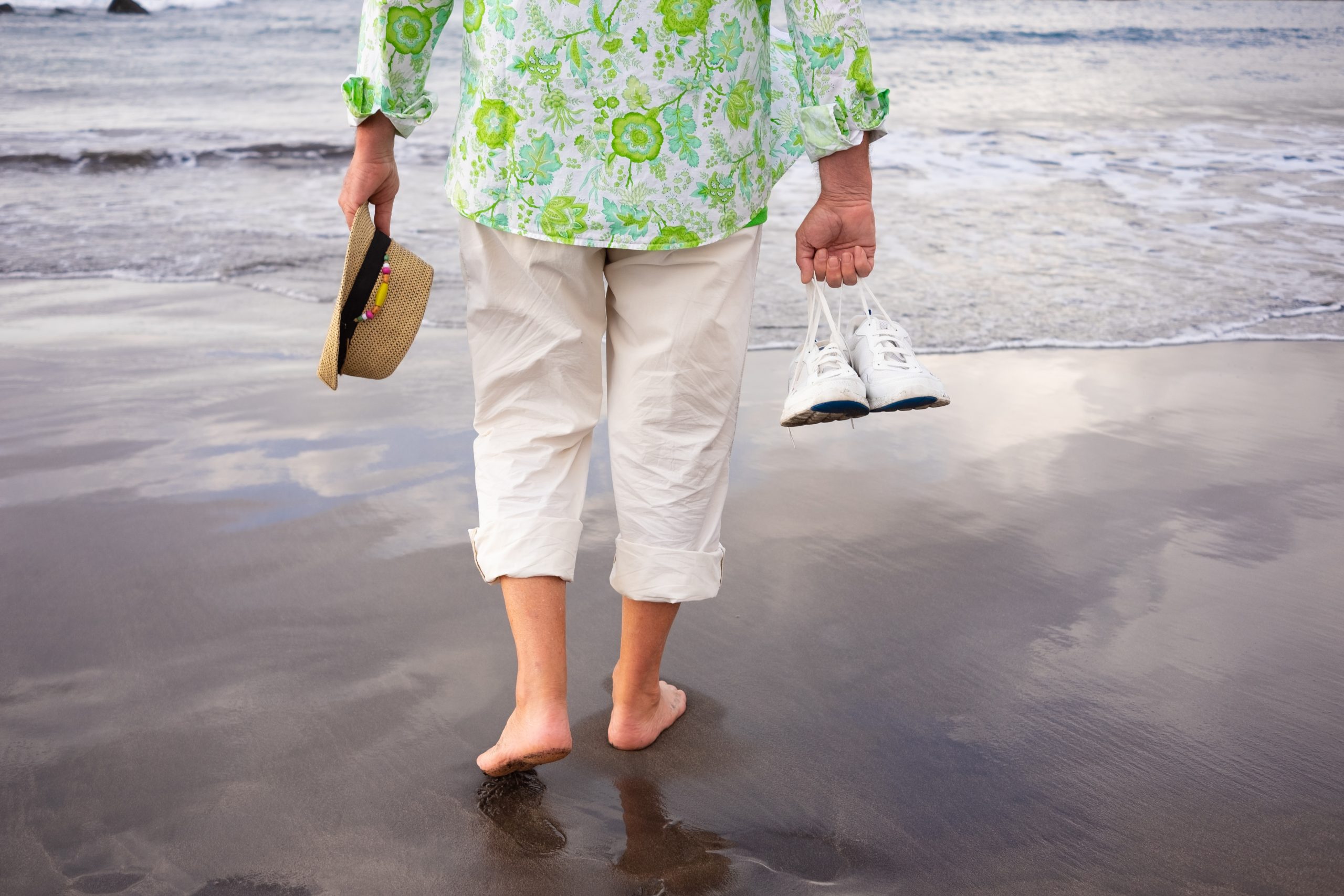 Back view of senior man walking on the beach barefoot holding sh